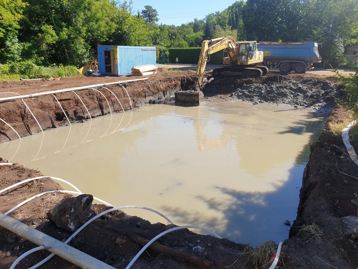 Zalany wodą wykop pod fundamenty na placu budowy, ilustrujący problem wysokiego poziomu wód gruntowych na działce. W tle pracuje koparka.